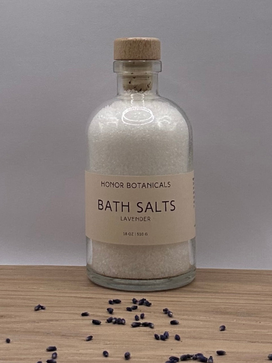Bottle of bath salts on a wooden surface with scattered lavender buds and gray background