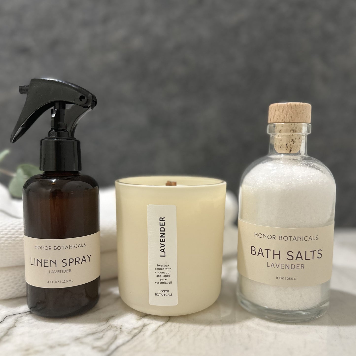 Honor Botanicals linen spray, lavender candle, and bath salts on a marble surface with a gray background.