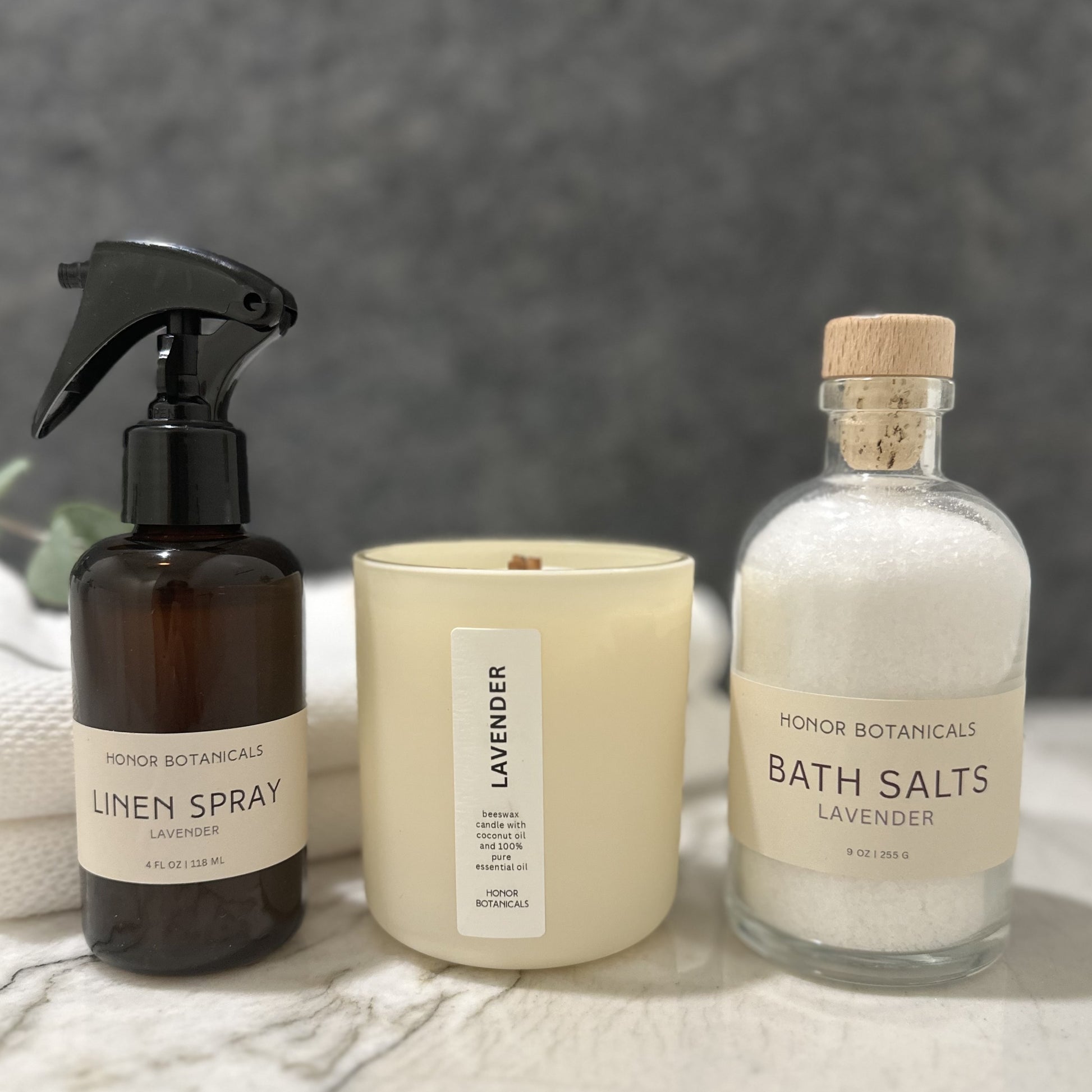Honor Botanicals linen spray, lavender candle, and bath salts on a marble surface with a gray background.